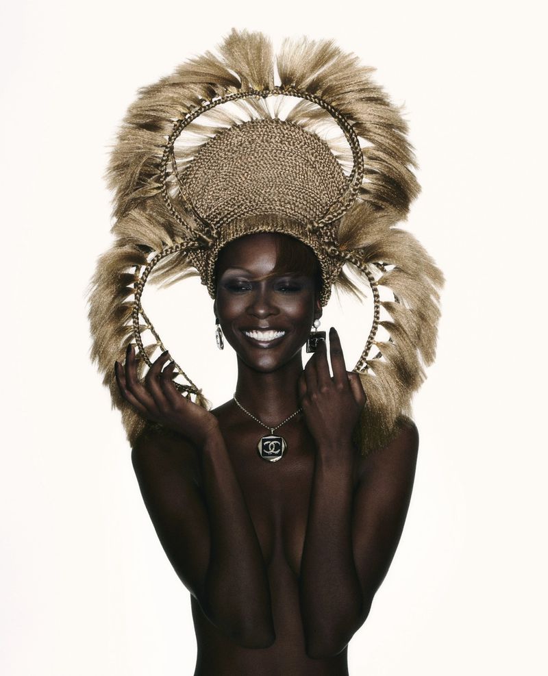 Portrait of a smiling Black woman with decorative gold headwear, jewelry, and bare shoulders against a white background in a studio setting.