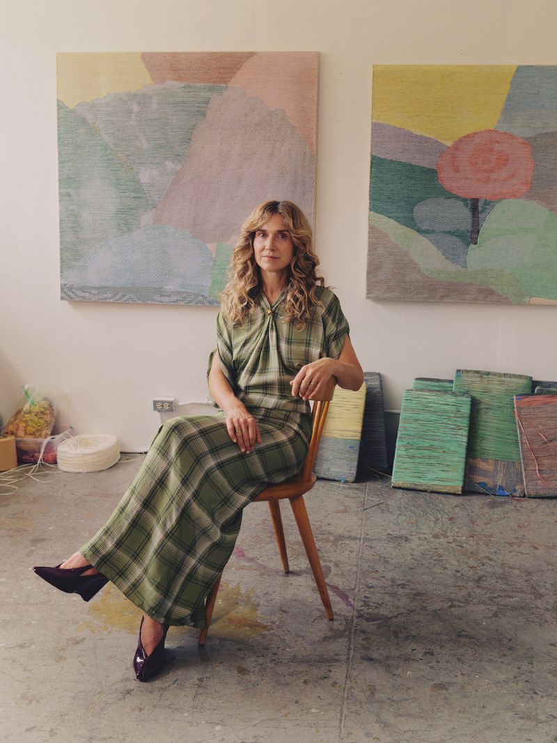 Woman in plaid dress sitting on chair in art studio with abstract paintings, creating a bright and artistic scene with natural light.