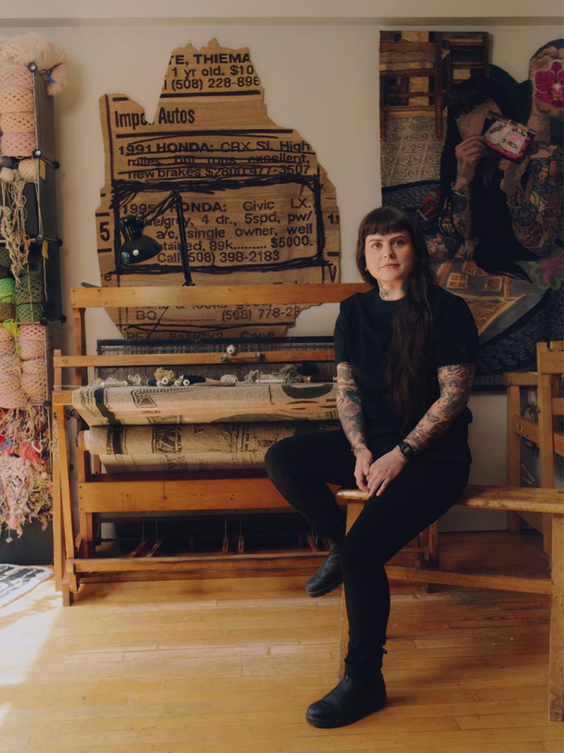 Adult woman sitting in front of weaving loom and wall art in an artist studio, with natural lighting indoors and black clothing.