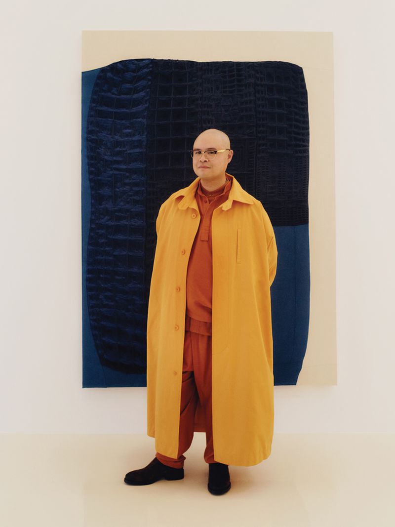Stylish adult with eyeglasses in a mustard overcoat, orange suit, and black shoes posing near a blue abstract painting indoors.