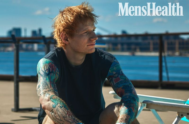 Red-haired man with tattoos sitting outdoors with cityscape and body of water background, wearing a dark shirt, featured in Mens Health.