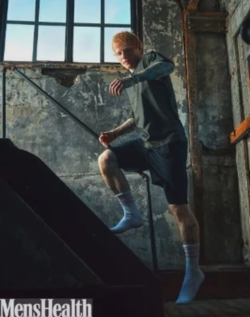 Ed Sheeran climbs industrial stairs in a gray T-shirt, navy shorts, and white socks, captured in medium shot for Men's Health.
