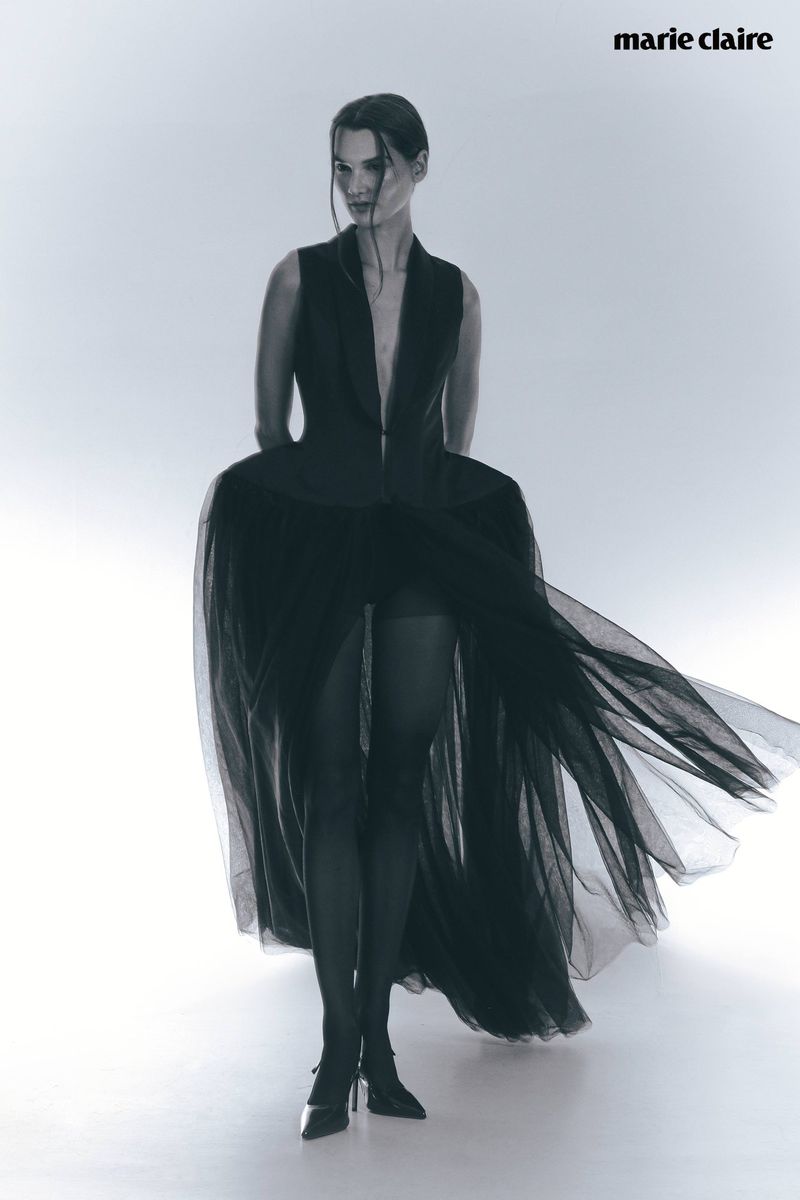 Fashion editorial shot of a woman in a dramatic black dress with a tulle skirt, posing in a photo studio with a white backdrop.