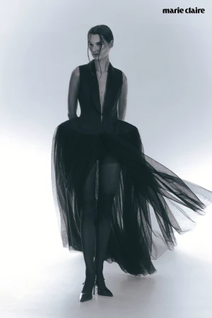 Fashion editorial shot of a woman in a dramatic black dress with a tulle skirt, posing in a photo studio with a white backdrop.