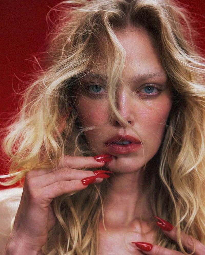 Close-up shot of a blonde adult woman with long, red stiletto nails posing against a vibrant red backdrop in a beauty style image.