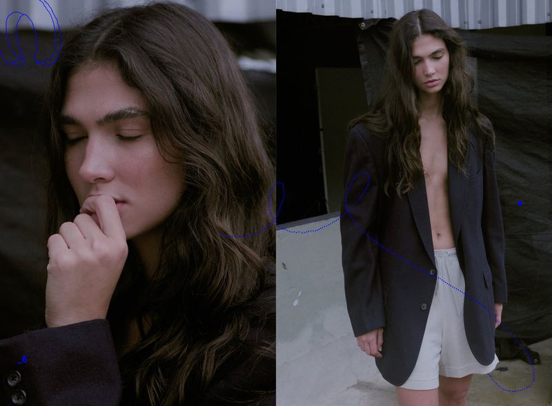 Portrait of a young person with long brown hair wearing a dark blazer and light shorts, posing in an outdoor urban environment.
