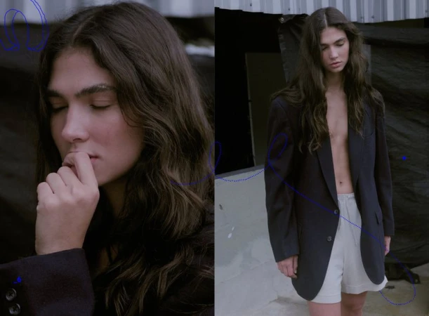 Portrait of a young person with long brown hair wearing a dark blazer and light shorts, posing in an outdoor urban environment.