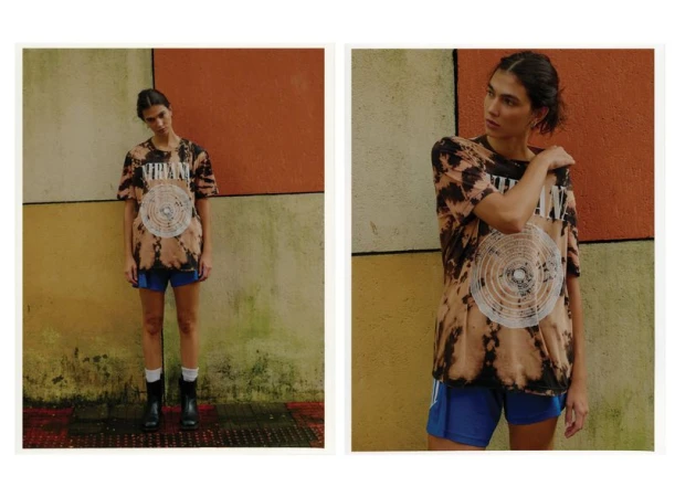 Woman in a 'Nirvana' tie-dye T-shirt and blue shorts stands against a multicolored wall, captured in an eye-level medium shot indoors.