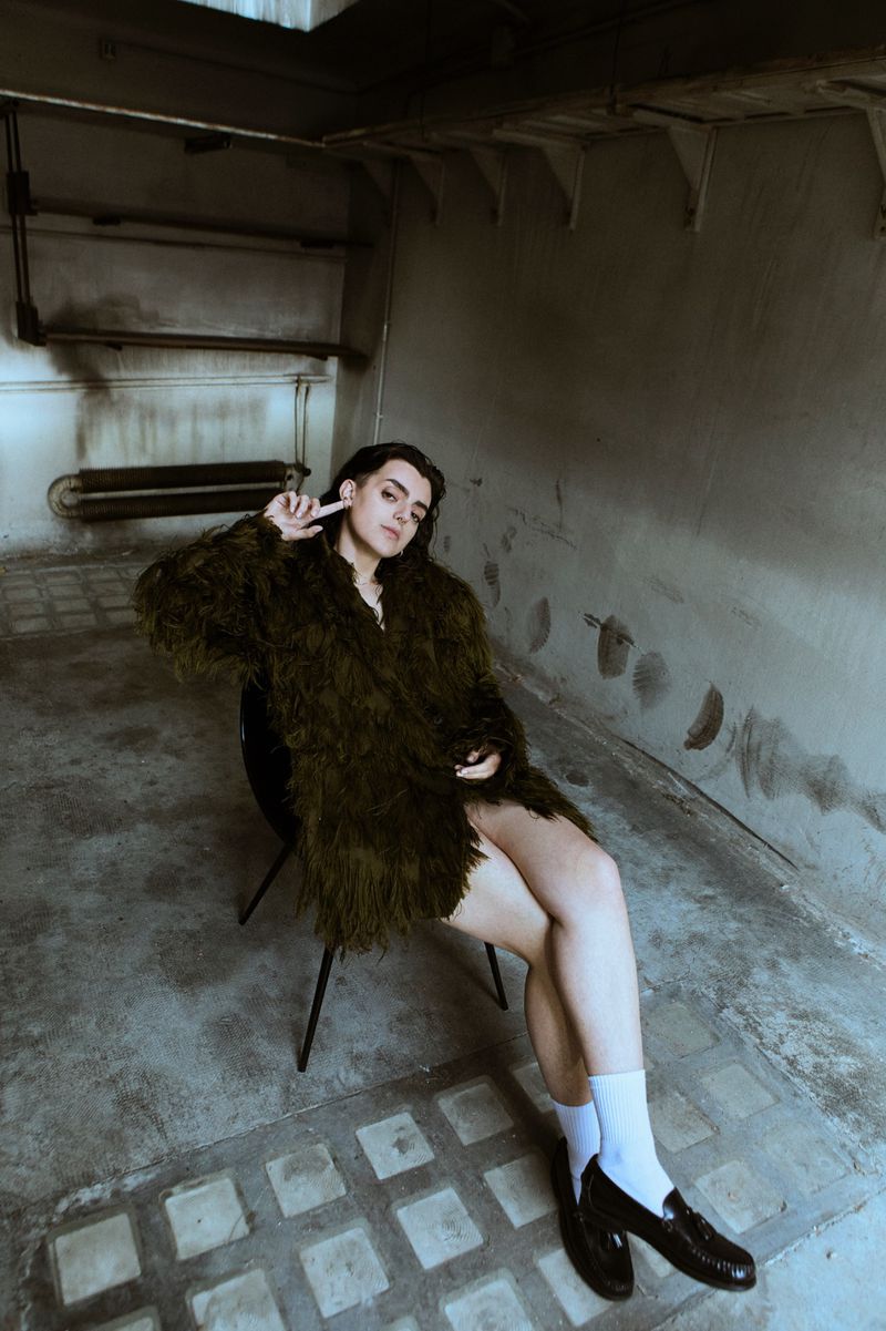 A woman sits on a chair indoors wearing a furry coat, white socks, and loafers with a concrete background and textured floor.
