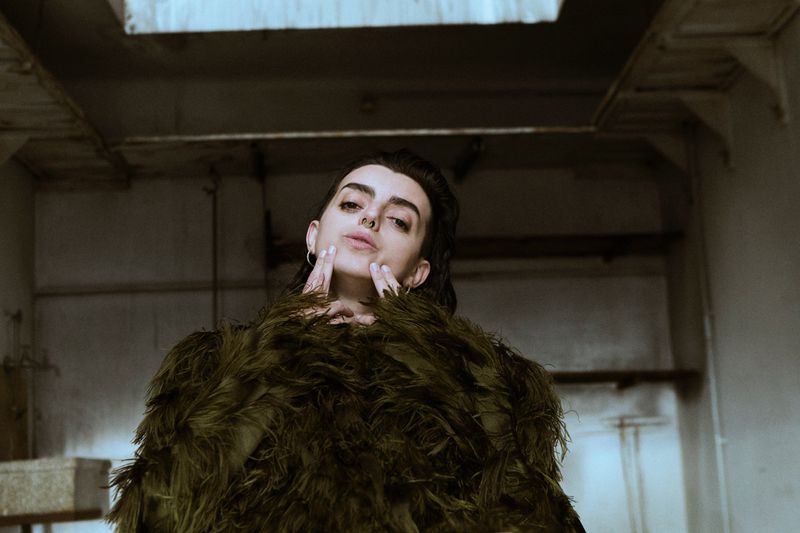Indoor portrait of a woman with dark hair in an olive green feather-like jacket, with her hands near her face looking at the camera.