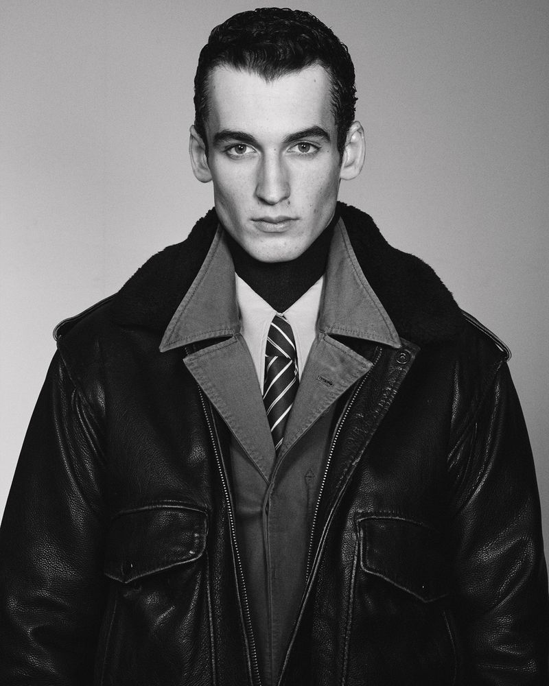 Portrait of a man wearing a dark leather jacket, layered collared shirt, and striped tie against a plain studio background.