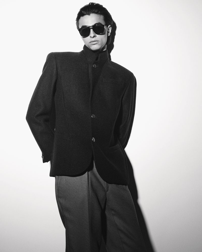 Black and white portrait of a male model wearing dark sunglasses and a structured jacket against a plain wall