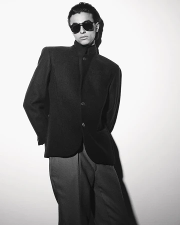 Black and white portrait of a male model wearing dark sunglasses and a structured jacket against a plain wall