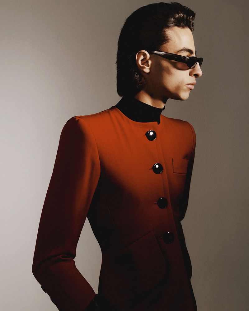 A profile view of a person with slicked-back hair, wearing narrow black sunglasses, a black turtleneck, and a fitted burnt orange buttoned jacket.