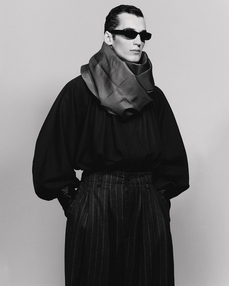 Black and white portrait of a male model with a scarf, wearing sunglasses, and a pinstripe skirt, posing in a photo studio.