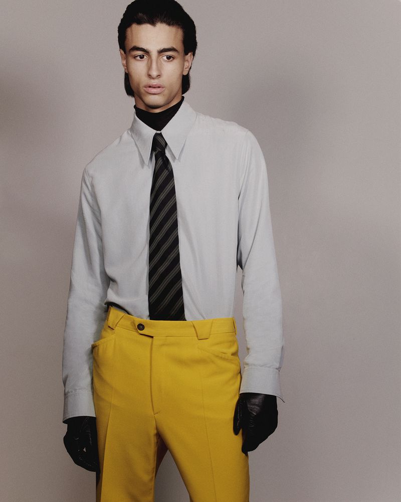 Fashion portrait of a young adult man in a gray shirt, striped tie, and yellow pants, standing against a gray backdrop.