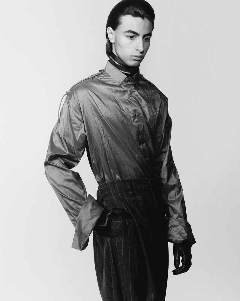 Monochrome portrait of a young adult with slicked-back hair in a satin shirt and pinstripe pants, standing indoors against a plain background.