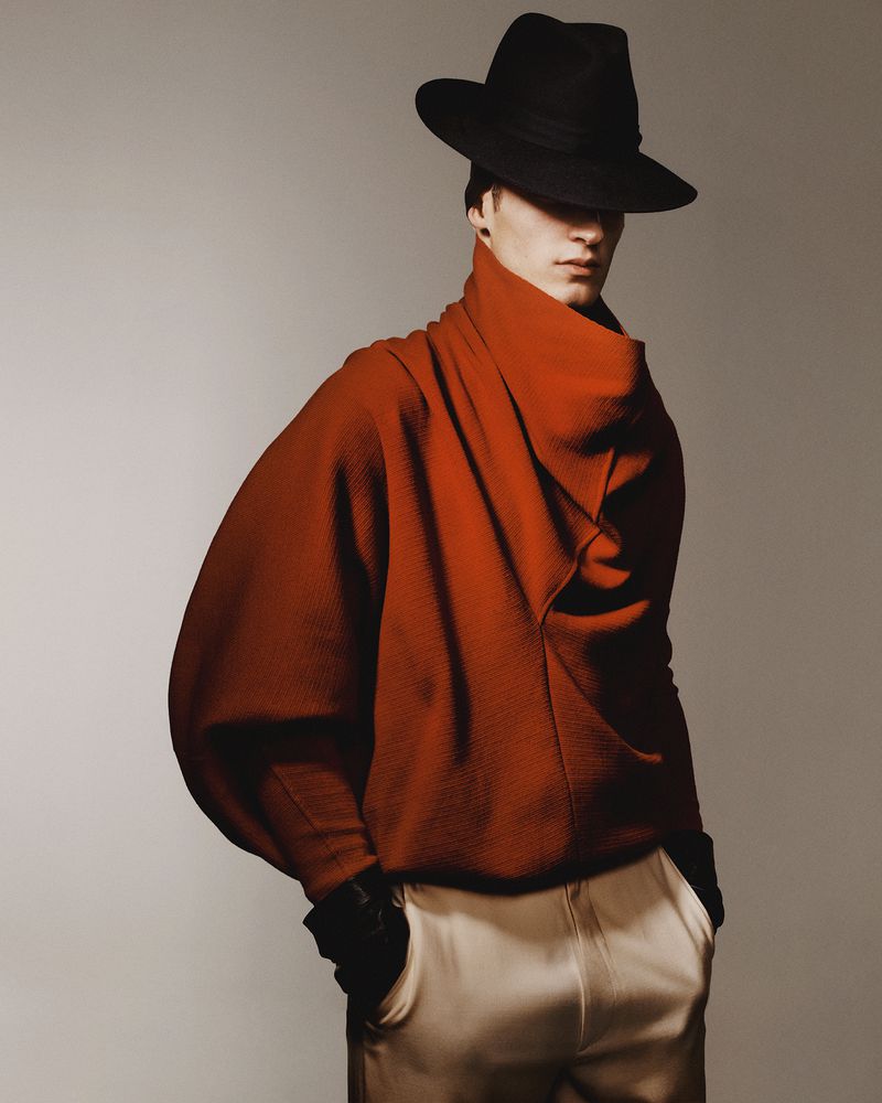Man in wide-brimmed hat and orange sweater posing in a studio with neutral background. Fashion editorial image with strong light and shadow.