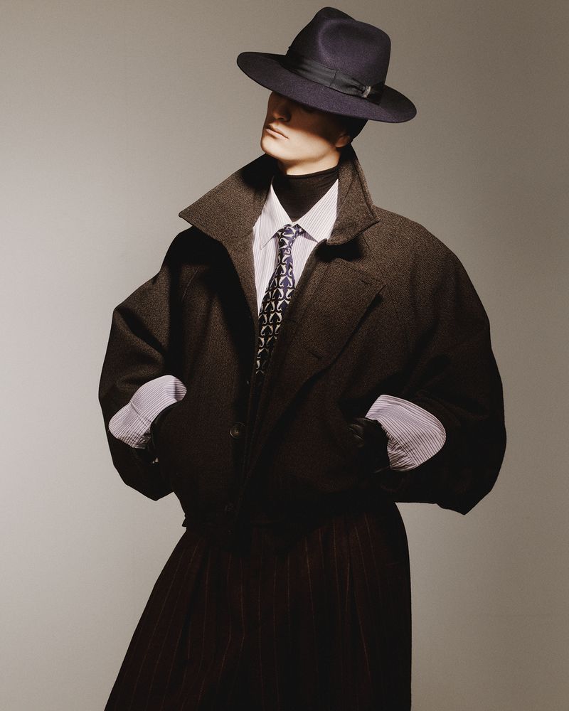 Stylish adult poses in oversized coat, dress shirt, tie, and fedora hat. Fashion model in studio against a grey backdrop.