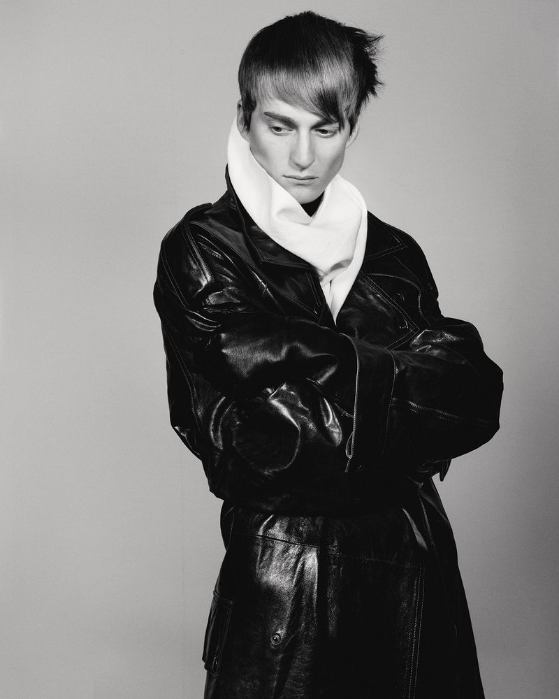 Portrait of a male model with dark hair, wearing a black leather trench coat and a white scarf, against a grey studio backdrop.