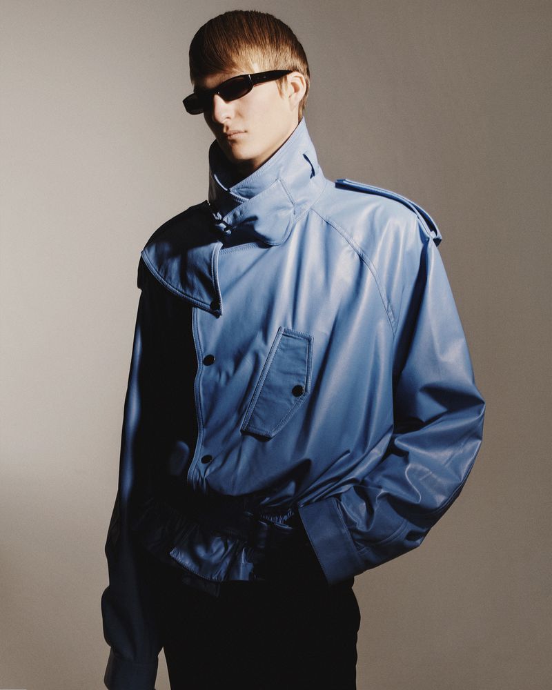 Fashionable man wearing a blue leather jacket and sunglasses posing in a studio. The lighting is soft, and the background is neutral.