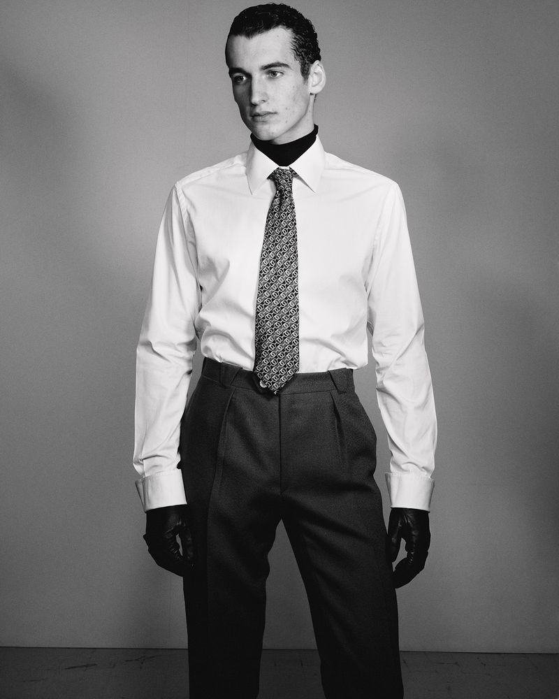 Studio portrait of a dark-haired male model in a white shirt and grey pants with a patterned tie and black leather gloves, indoors.