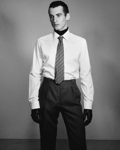 Studio portrait of a dark-haired male model in a white shirt and grey pants with a patterned tie and black leather gloves, indoors.