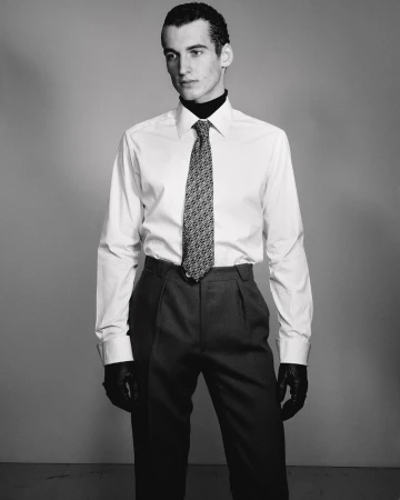 Studio portrait of a dark-haired male model in a white shirt and grey pants with a patterned tie and black leather gloves, indoors.
