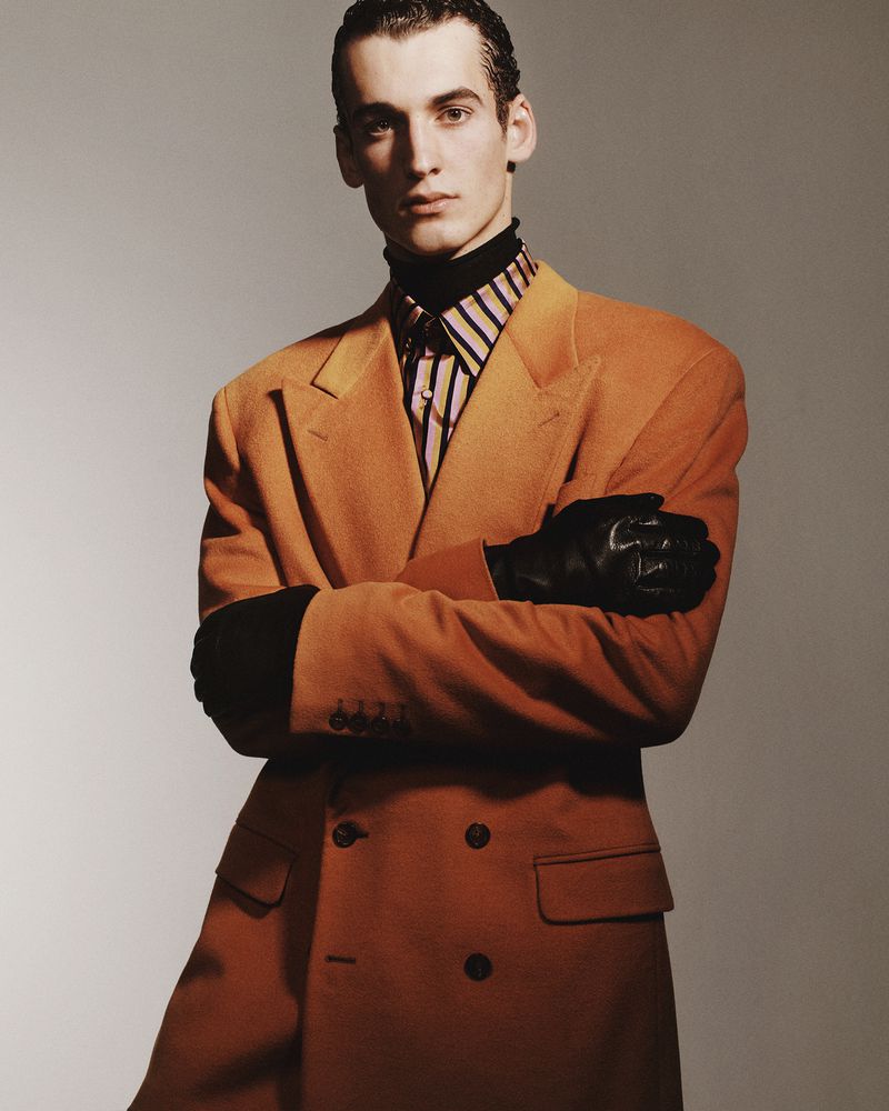 Male model with arms crossed wearing an orange double-breasted coat and leather gloves against a grey background.