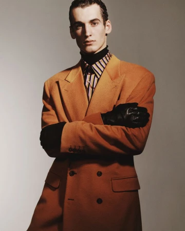 Male model with arms crossed wearing an orange double-breasted coat and leather gloves against a grey background.