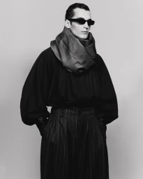 Model wearing dark sunglasses, voluminous draped neck scarf, and pinstripe trousers against a plain studio background.