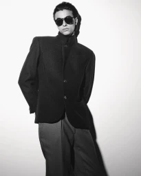 Black and white portrait of a male model wearing dark sunglasses and a structured jacket against a plain wall