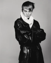 A young male model with dark hair wearing a black leather coat and a white high-neck collar against a plain grey background