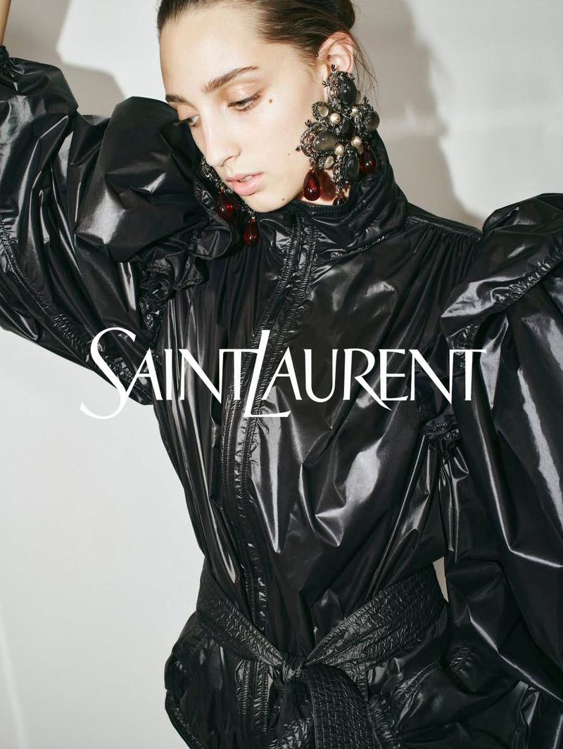 Fashion shot of a woman in a black, shiny jumpsuit with large, jeweled earrings, posing against a plain background. Saint Laurent logo visible.