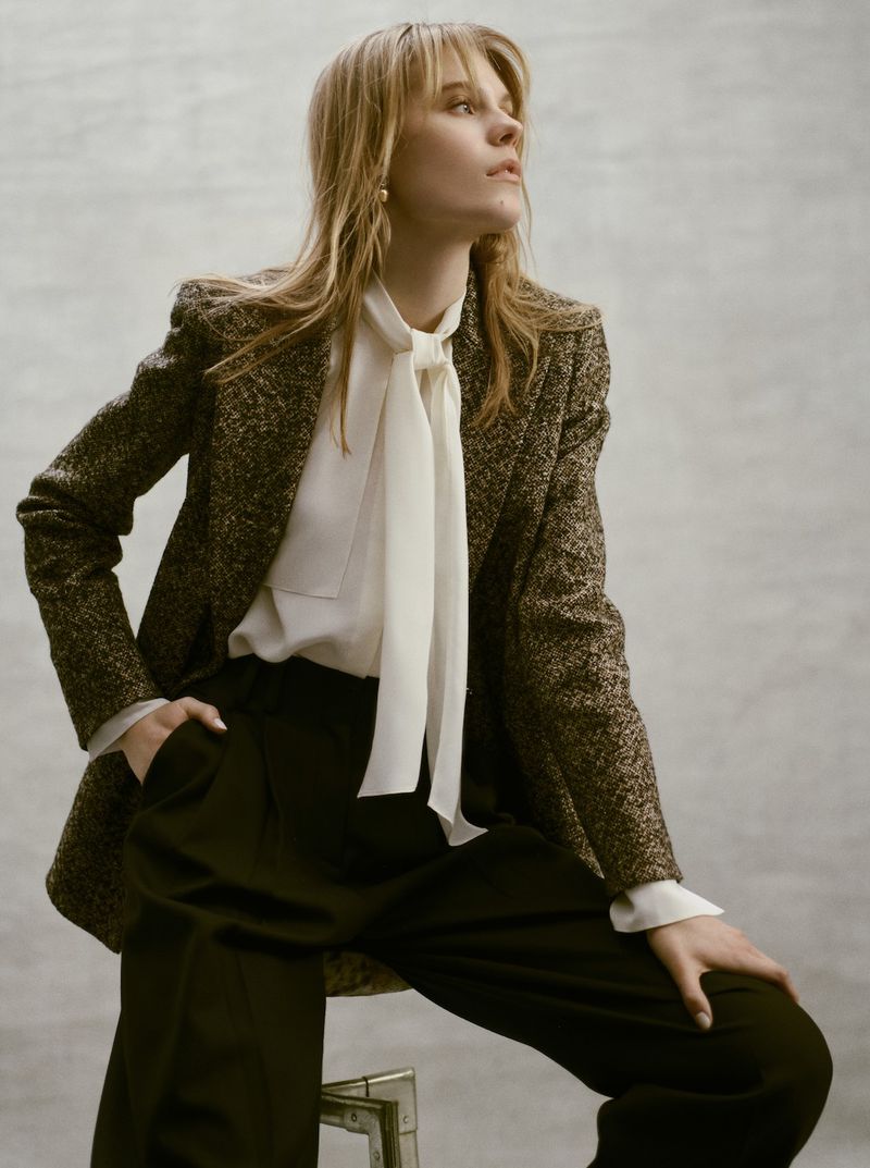 Blonde woman poses in a tweed blazer, blouse with a tie, and black slacks, gazing off-camera. Fashion portrait in a studio setting.