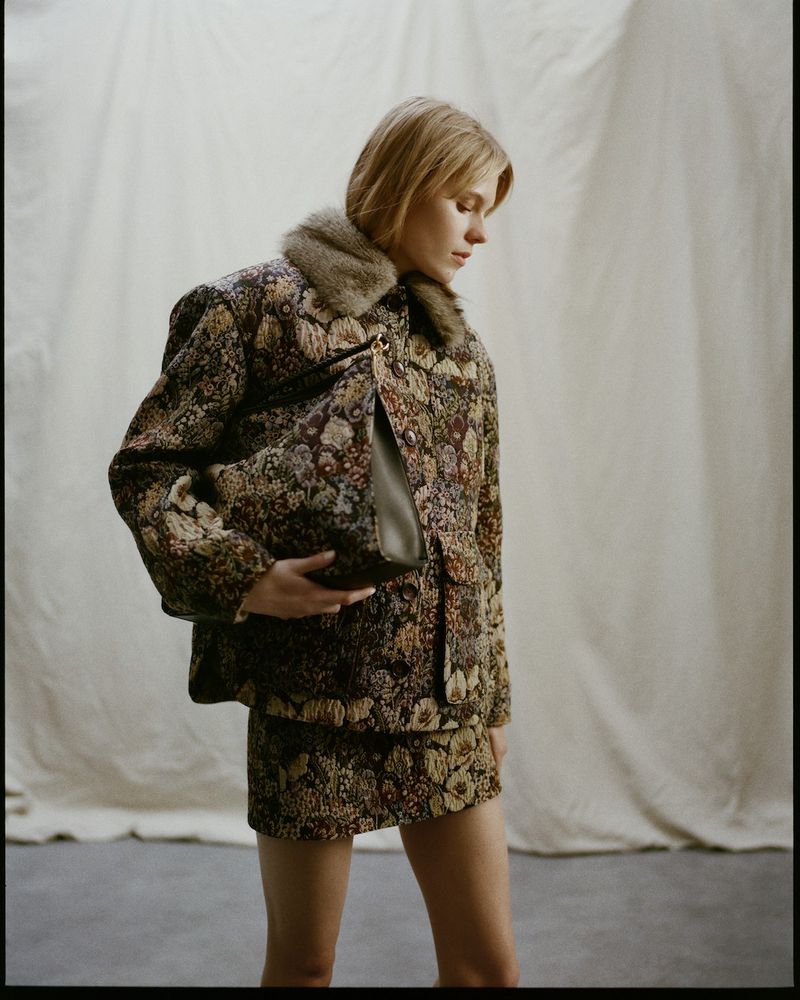 Blonde woman in a floral patterned jacket with a fur collar and matching skirt holding a purse, against a light beige background.