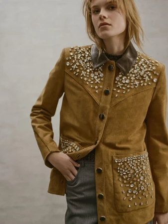 Blonde woman posing in a tan suede jacket embellished with rhinestones, with a dark leather collar and paired with grey trousers.
