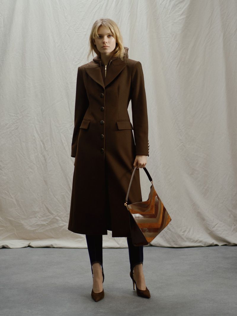 Full-length shot of a blond adult female in a long brown coat holding a handbag, posing in front of a white muslin backdrop.