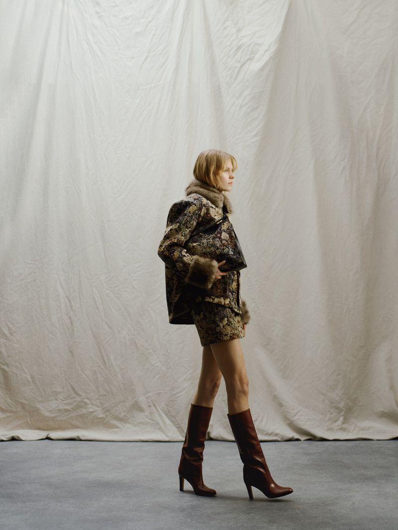 Elegant adult woman in matching print jacket and mini skirt with brown boots posing against a draped off-white fabric backdrop.