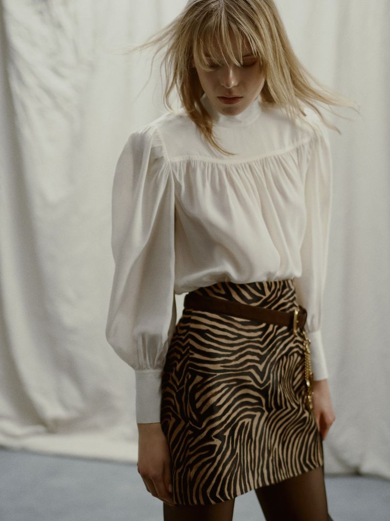 A woman poses in a white blouse and zebra print skirt in front of a white curtain, creating a fashionable studio portrait.