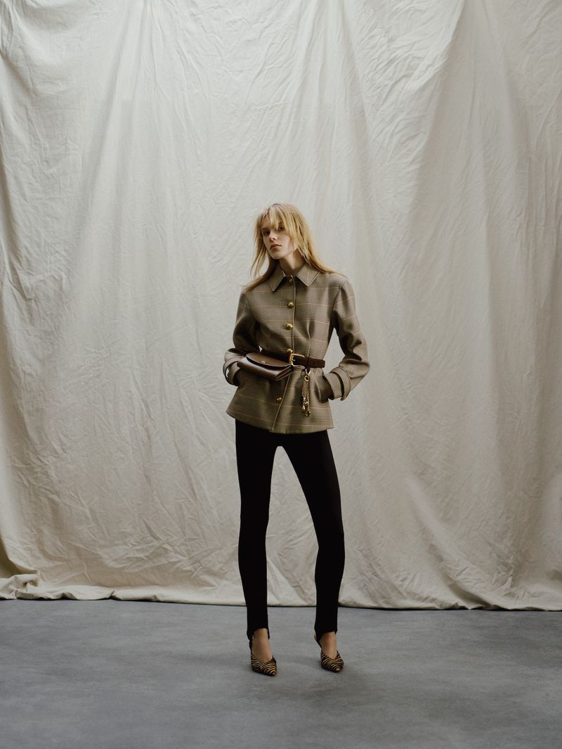Full-length shot of a woman in a brown jacket and black leggings, posing against a neutral backdrop, showcasing fashion and style.
