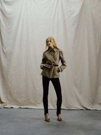 Full-length shot of a woman in a brown jacket and black leggings, posing against a neutral backdrop, showcasing fashion and style.