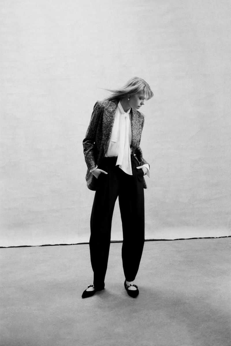 Fashion editorial of a woman in a tweed blazer, white blouse, and black trousers. She poses in a photo studio with a neutral background.