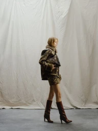 Model wearing a patterned jacket and skirt with knee-high boots against a draped fabric background