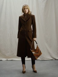 Model wearing a structured brown coat and holding a patterned handbag against a draped fabric backdrop.