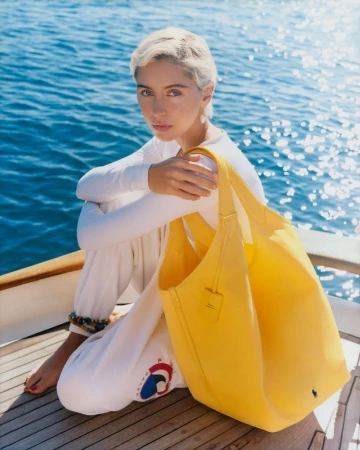 Blonde woman sits on boat deck with yellow tote bag against blue ocean backdrop on sunny day, wearing white clothing. Fashion shot.
