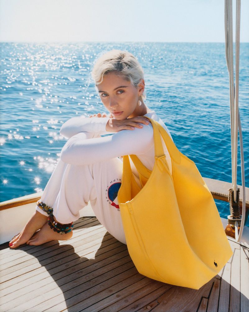 Young woman with short blonde hair in white outfit sits on a yacht deck with a bright yellow tote bag against an ocean backdrop.