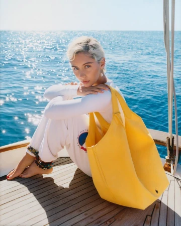 Young woman with short blonde hair in white outfit sits on a yacht deck with a bright yellow tote bag against an ocean backdrop.