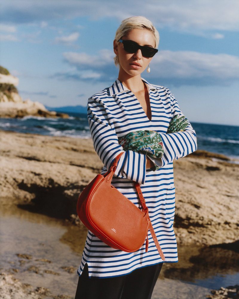 A woman in a striped shirt and sunglasses poses with a red handbag on a rocky beach with a blue sea and sky in the background.