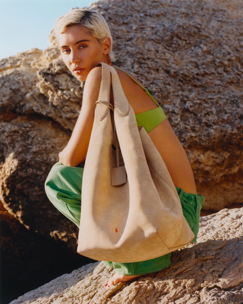 Woman poses outdoors on rocks with neutral tote bag, green pants and top, blonde pixie cut, sunlit. Fashion or lifestyle imagery.